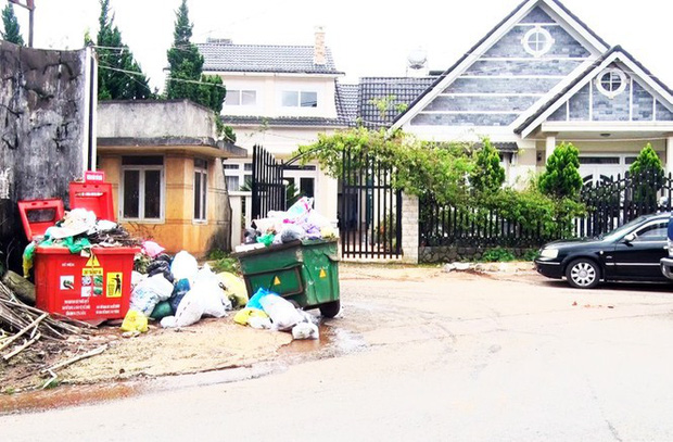 Thùng rác màu cam và màu xanh đặt cạnh nhau và đều đầy ắp rác.