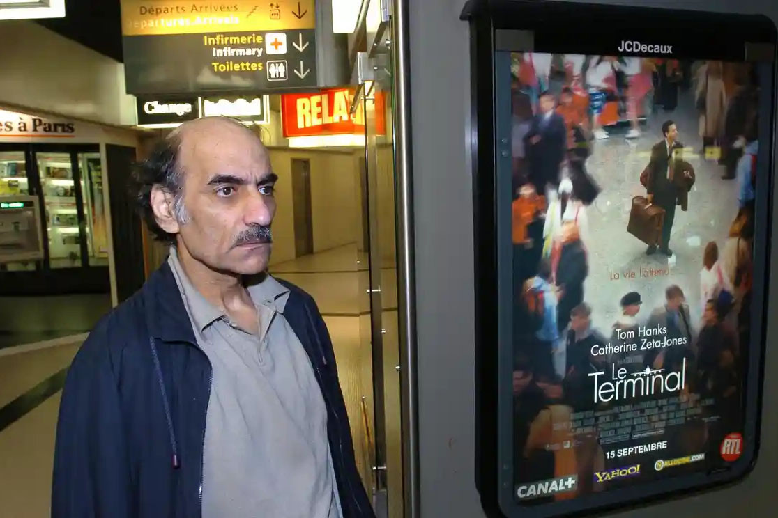 Ông Nasseri bên tấm biển quảng cáo về bộ phim “The Terminal” tại nhà ga số 1 của sân bay Charles De Gaulle, Paris, Pháp năm 2004. Ảnh: The Guardian