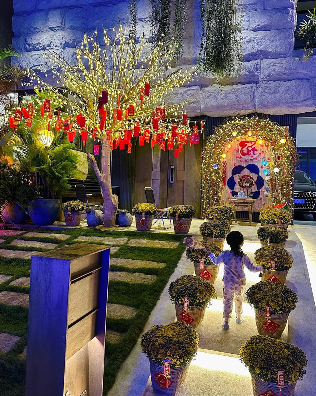 Lối vào nhà rực vàng bởi đèn và các chậu cúc xếp đều nhau. Bé Suchin vừa được mẹ chụp bộ ảnh mới trước nhà. Bên trong, vợ chồng Quốc Cường - Đàm Thu Trang trồng một cây mai lớn.